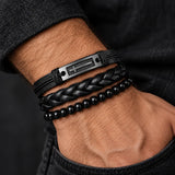 Man wearing a SEYOR three-piece black bracelet set with a cross plate, thick braided leather band and polished black beads.