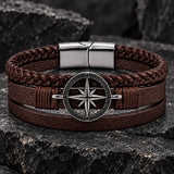 Close-up of a men’s brown braided leather bracelet featuring a silver-tone compass centerpiece on a dark stone background.