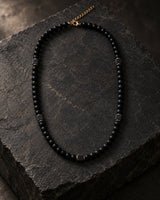 Matte black onyx beaded necklace with square lava stone accents and gold-tone details displayed on rugged dark stone.