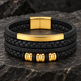 Close-up of a SEYOR men’s multilayer black leather bracelet featuring braided bands, a polished gold-tone bar, gold spacers and magnetic clasp.