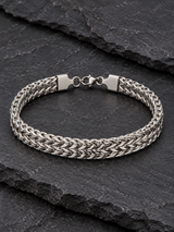 Men’s wide silver-tone woven chain bracelet with a lobster clasp displayed on rugged black stone.